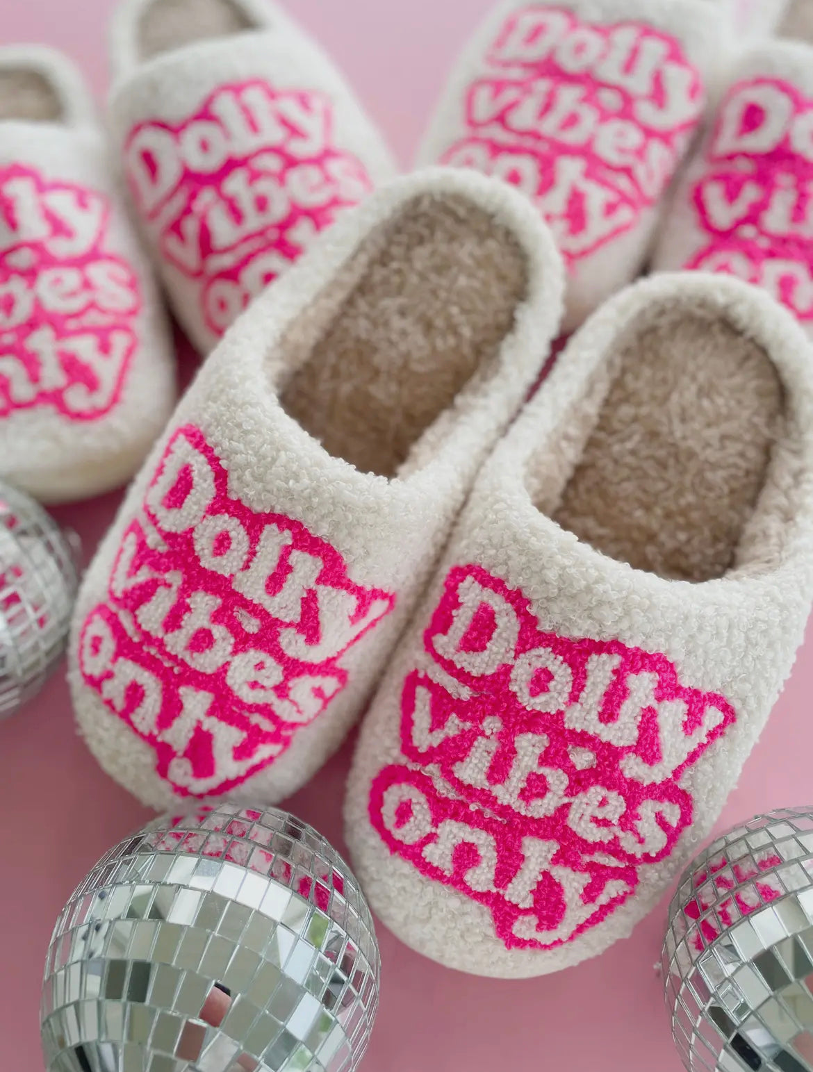 A pair of white slippers with 'Dolly Vibes Only' written in pink text on the top, displayed among silver disco balls.