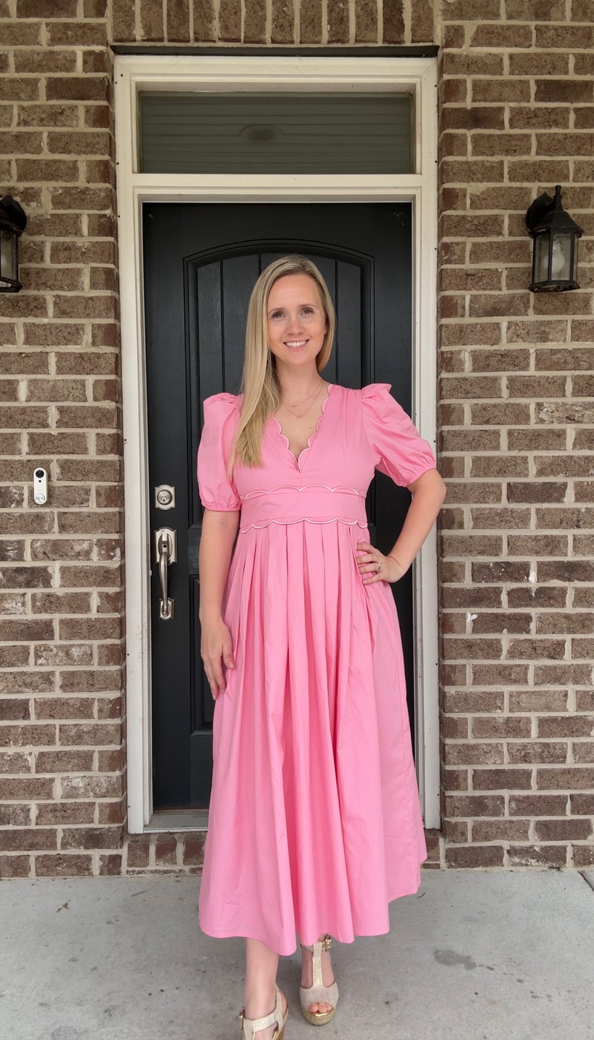 A person wearing a pink scallop midi dress with a v-neckline, scalloped details, and puff sleeves. The dress has a belted waist and a concealed zipper.